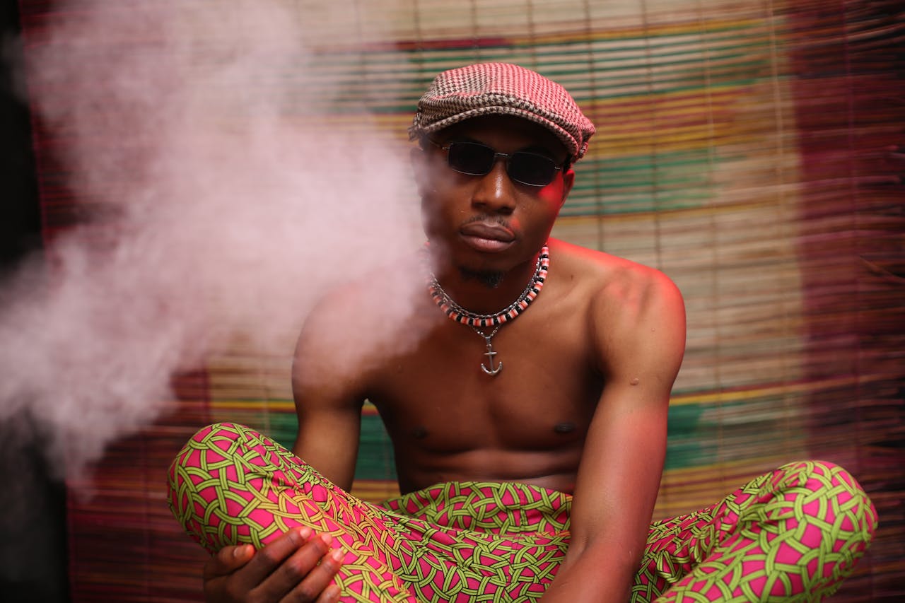 mobile-02 Shirtless black guy in colorful pants and cap sitting in dark room and smoking cannabis